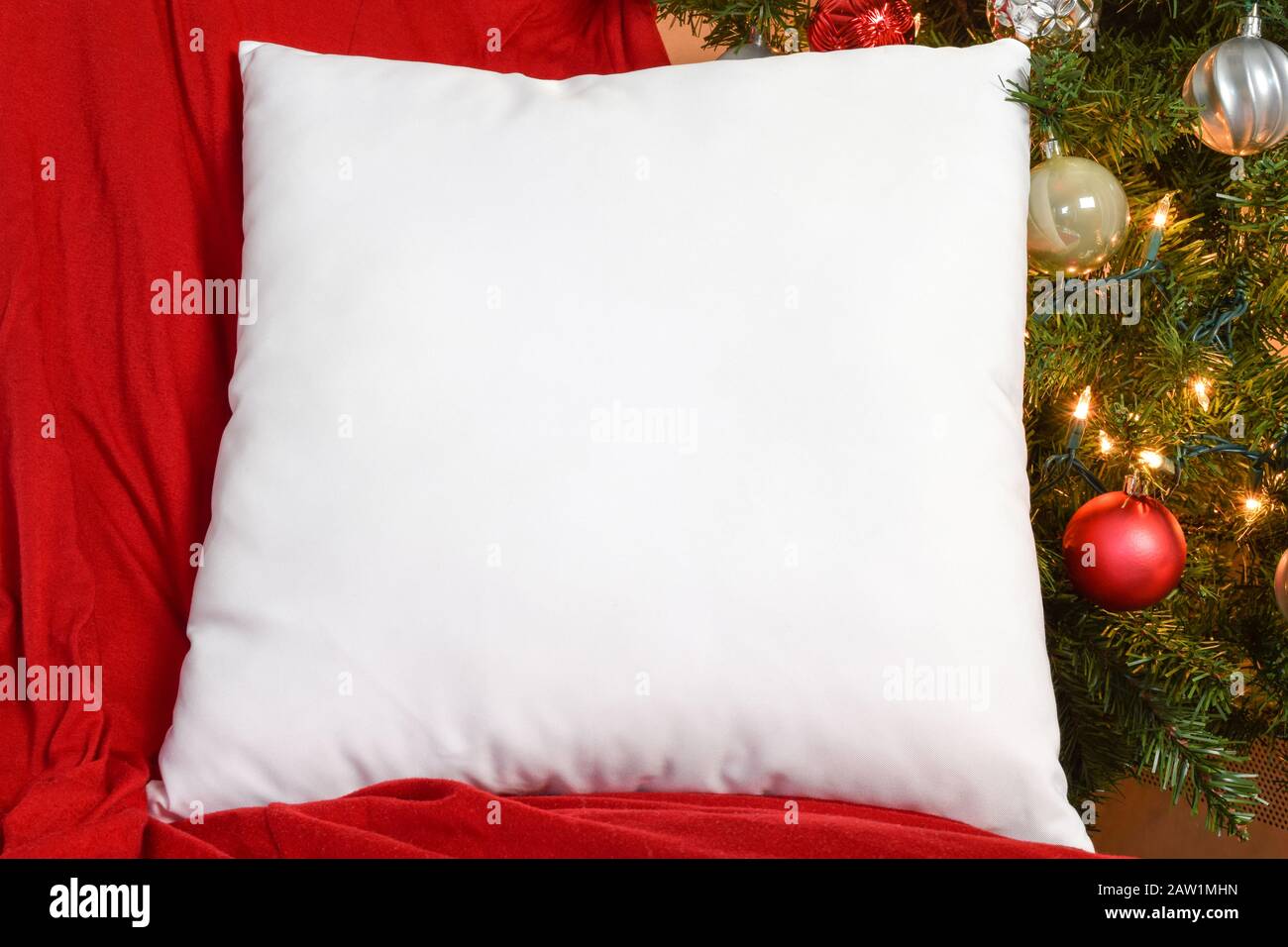 White Square PIllow Mockup Resting on Red Blanket with Lit Up Christmas Tree in Background Stock Photo