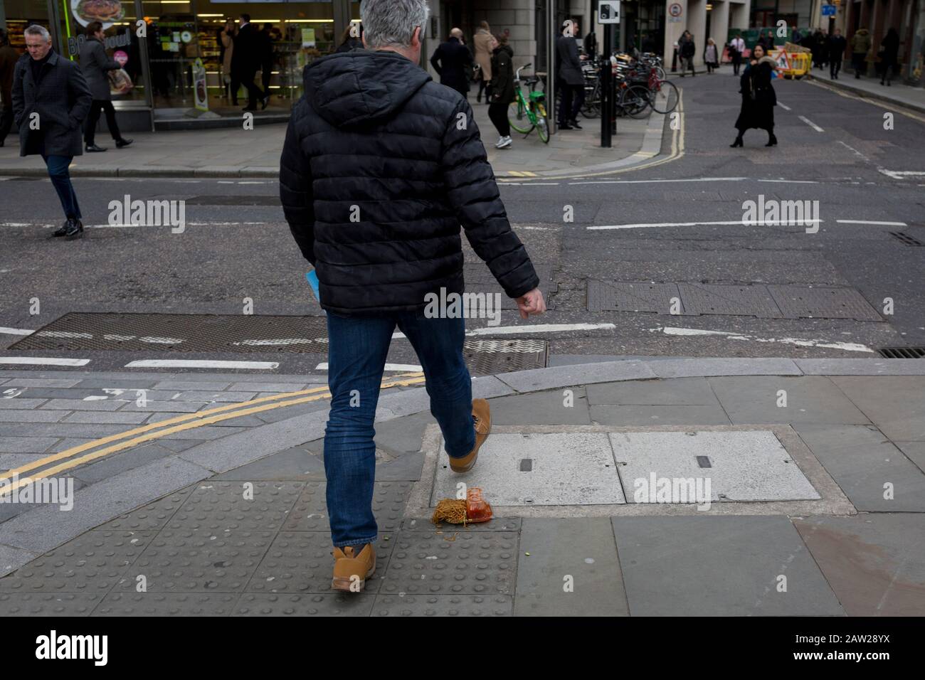 Pedestrians avoid a noodle and sauce takeaway, dropped and discarded on the pavement during lunch-hour in the capital's financial district, on 4th February 2020, in the City of London, England. The lunchtime meal was being carried along the street when its heat and moisture made it drop through the bottom of a paper bag, turning it upside down and lying perfectly on the pavement as city workers emerged from their offices. Those who saw it in time stepped over the greasy obstacle but the distracted (mostly by walking with phones to ears), stepped in it and helping spread it across the pavement. Stock Photo