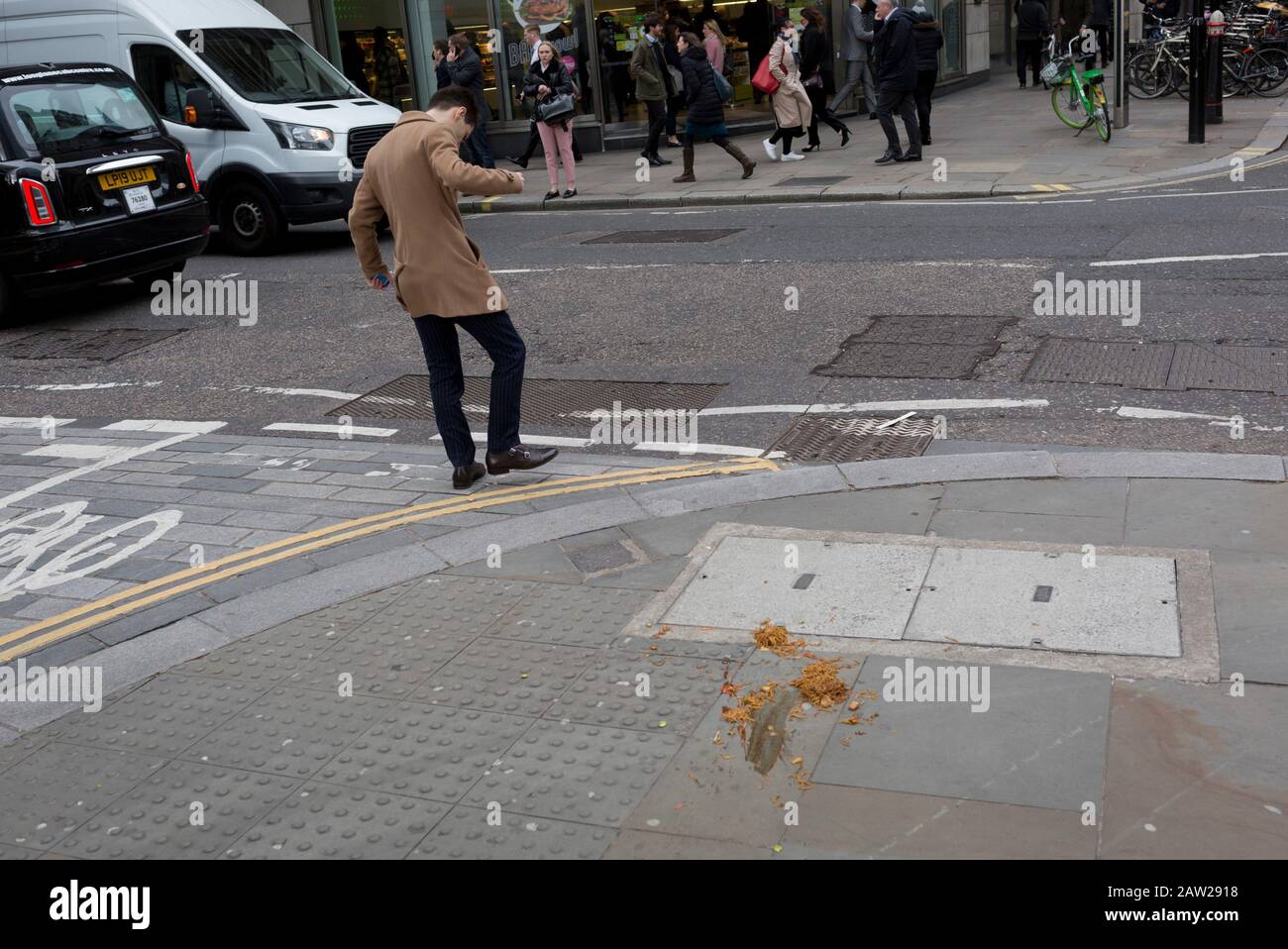 A pedestrian fails to avoid a noodle and sauce takeaway, dropped and discarded on the pavement during lunch-hour in the capital's financial district, on 4th February 2020, in the City of London, England. The lunchtime meal was being carried along the street when its heat and moisture made it drop through the bottom of a paper bag, turning it upside down and lying perfectly on the pavement as city workers emerged from their offices. Those who saw it in time stepped over the greasy obstacle but the distracted (mostly by walking with phones to ears), stepped in it and helping spread it across the Stock Photo