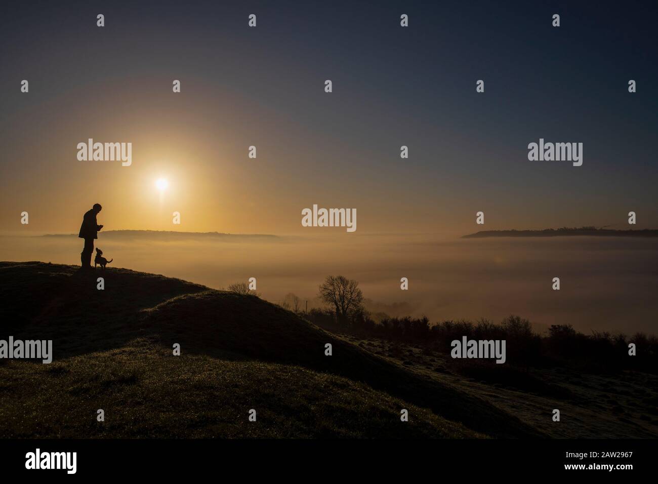 A man and his dog admire the cloud inversion over the City of Bath in Somerset at sunrise from Solsbury Hill on a winter's day. Stock Photo