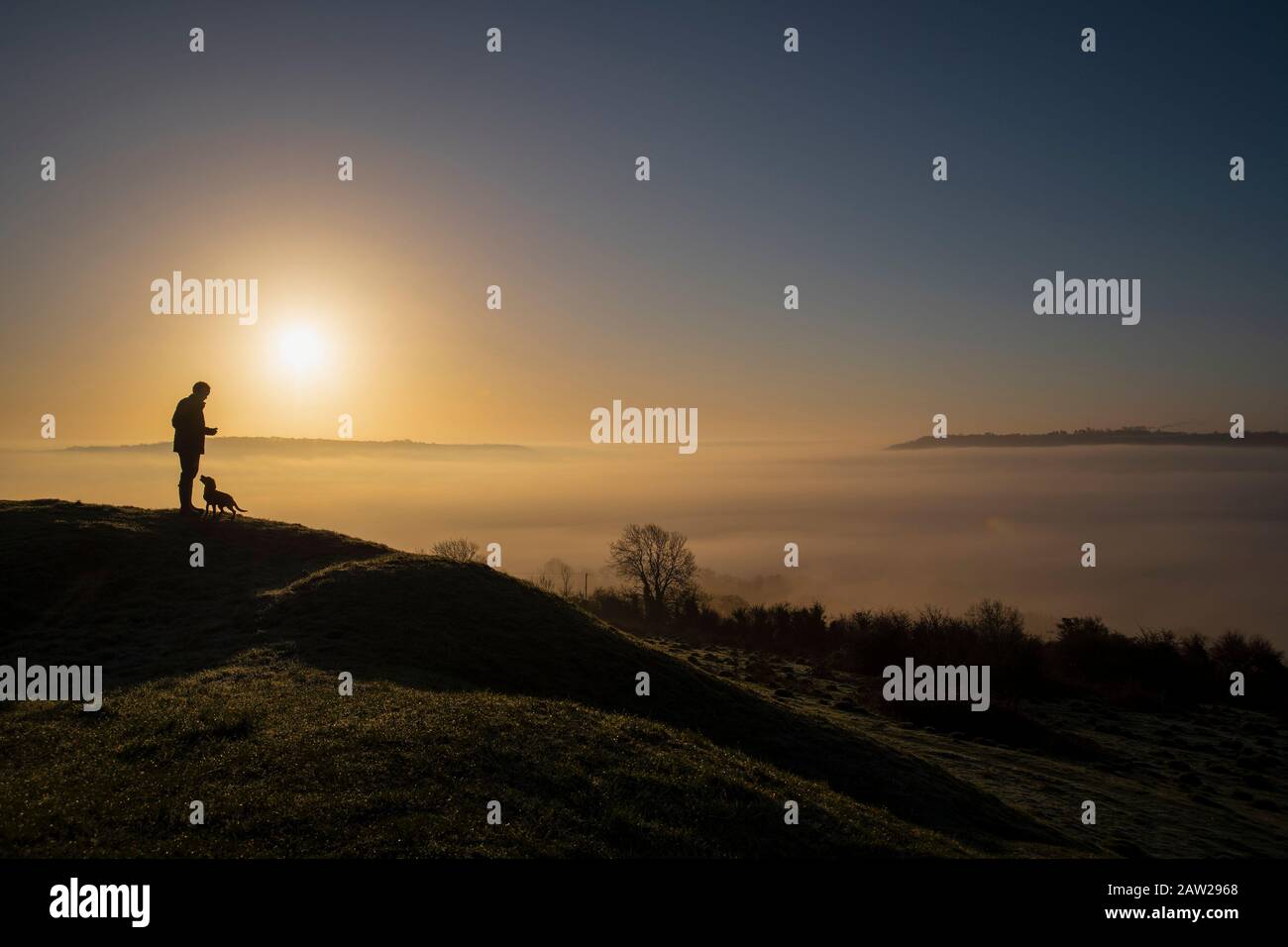 A man and his dog admire the cloud inversion over the City of Bath in Somerset at sunrise from Solsbury Hill on a winter's day. Stock Photo