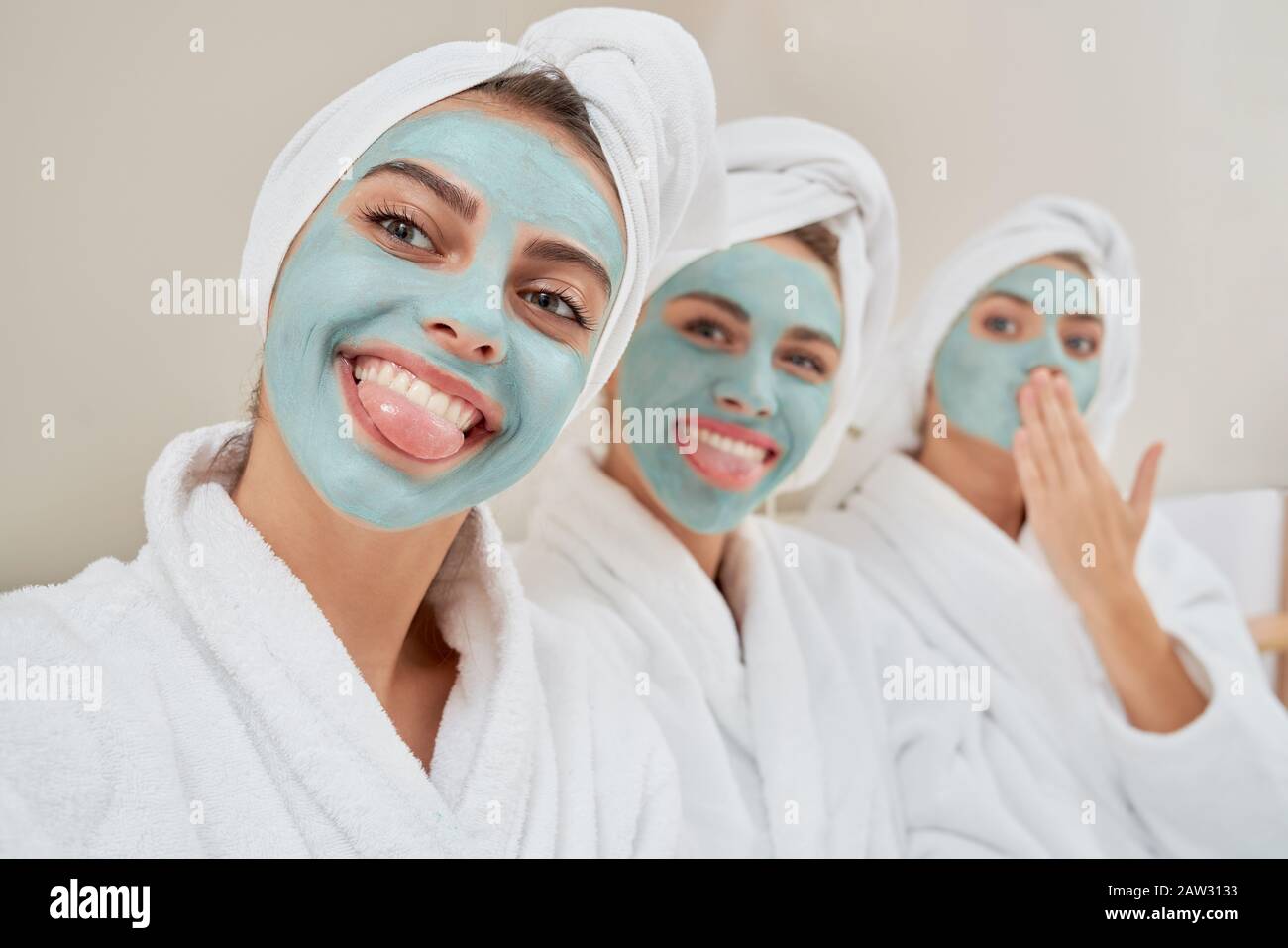 Close up of smiling group of girls with cosmetic masks, in towels on heads and bathrobes taking selfie after shower in bedroom. Three emotional girls having fun. Concept of friendship, skincare. Stock Photo