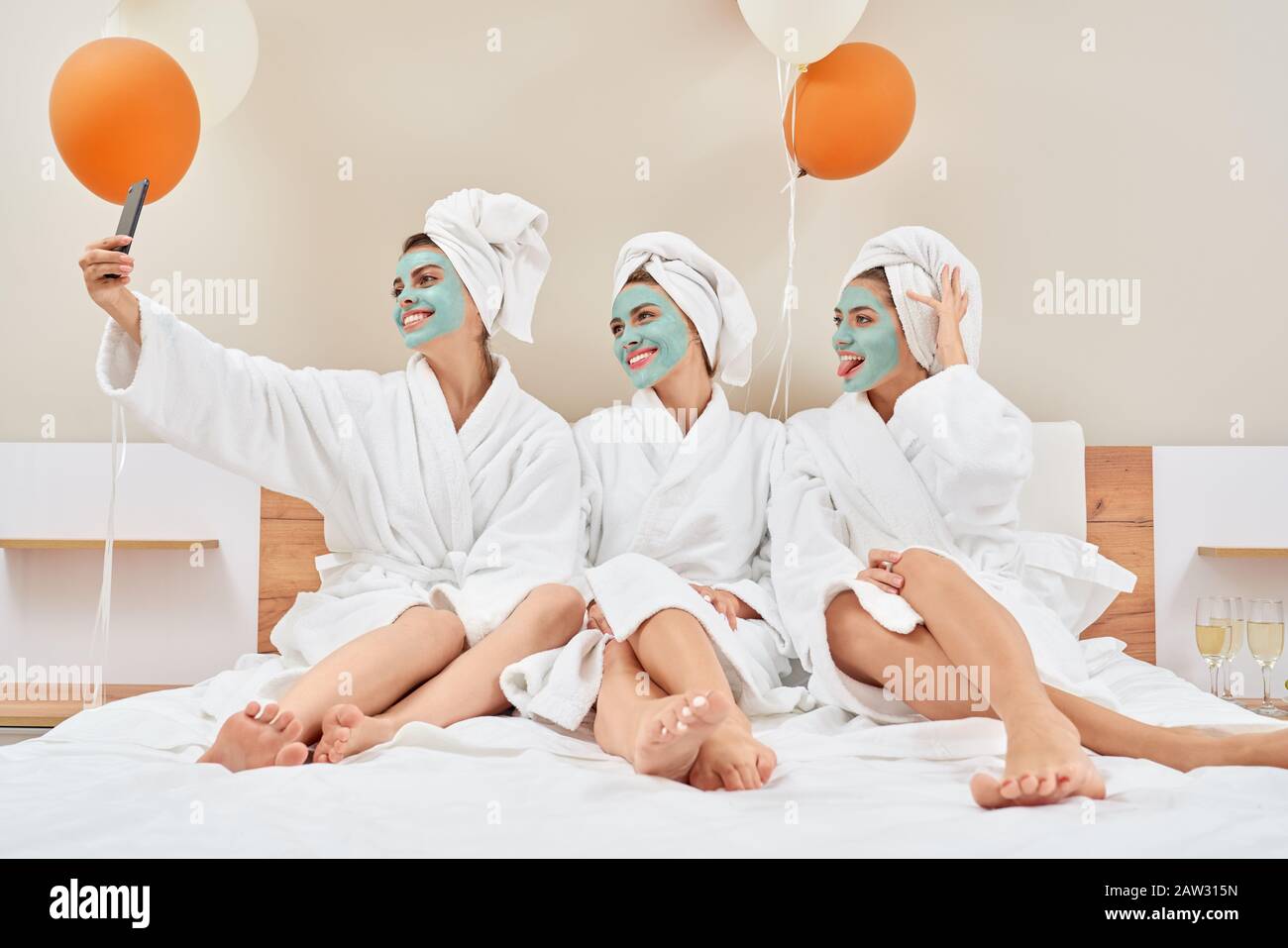 Front view of smiling group of girls with cosmetic masks, in towels on heads and bathrobes having fun at home. Three pretty female friends taking selfie in bedroom with balloons, champagne nearby. Stock Photo