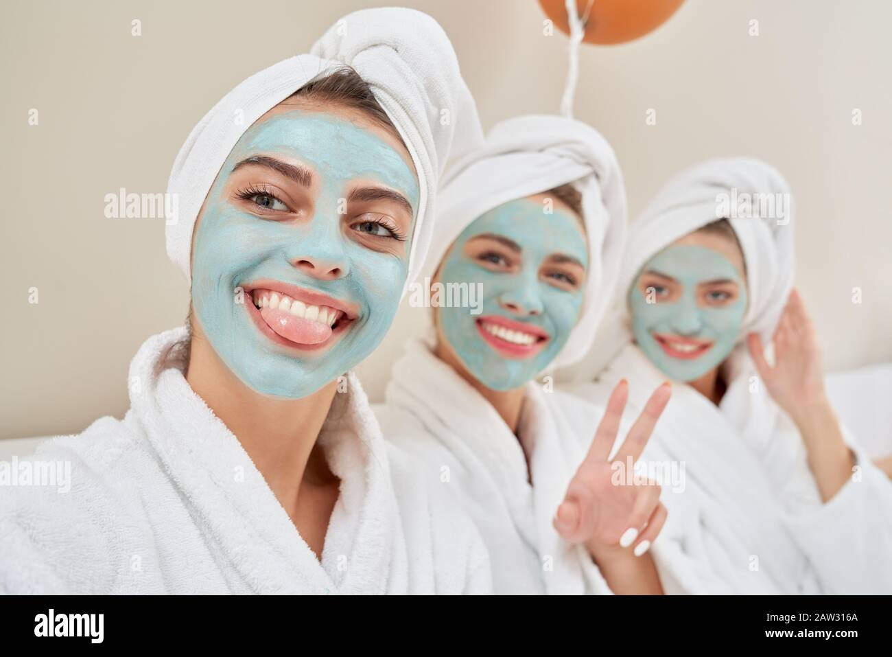 Close up of smiling group of girls with cosmetic masks, in towels on heads and bathrobes having fun at home. Three pretty girls taking selfie after shower in bedroom. Concept of friendship, skincare. Stock Photo