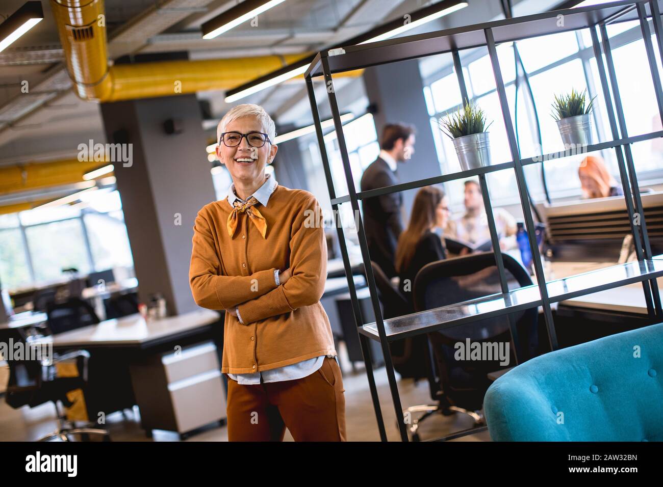Portrait of smiling senior woman standing outside meeting room with team discussing work in background Stock Photo