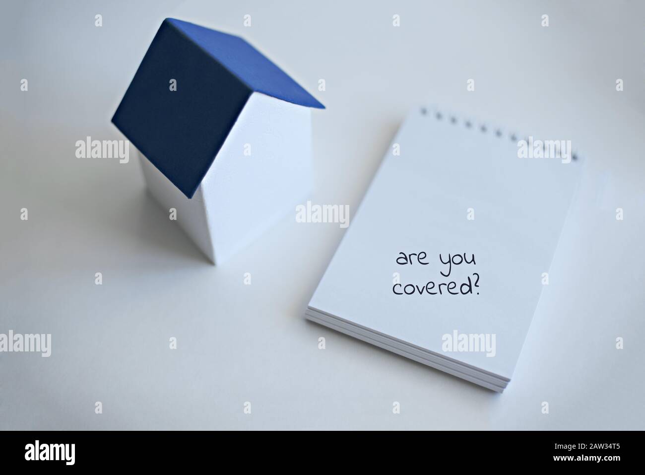 A layout of a house with a classic blue roof stands on a white surface next to the notebook with text Are you covered. Real estate insurance concept. Stock Photo