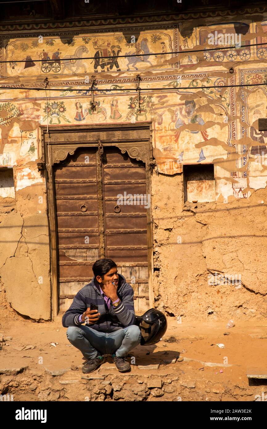 India, Rajasthan, Shekhawati, Mandawa, Akram Ka Haveli, man squatting below remnants of historic old painted walls with old wooden door Stock Photo