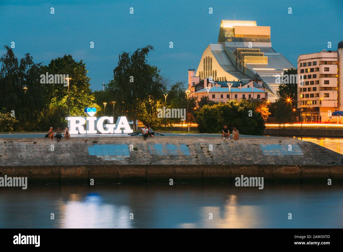 Riga, Latvia - June 30, 2016: Sitting People On Edge Of Urban Concrete Embankment Of Daugava River Around The Glowing City Name Sign In Summer Evening Stock Photo