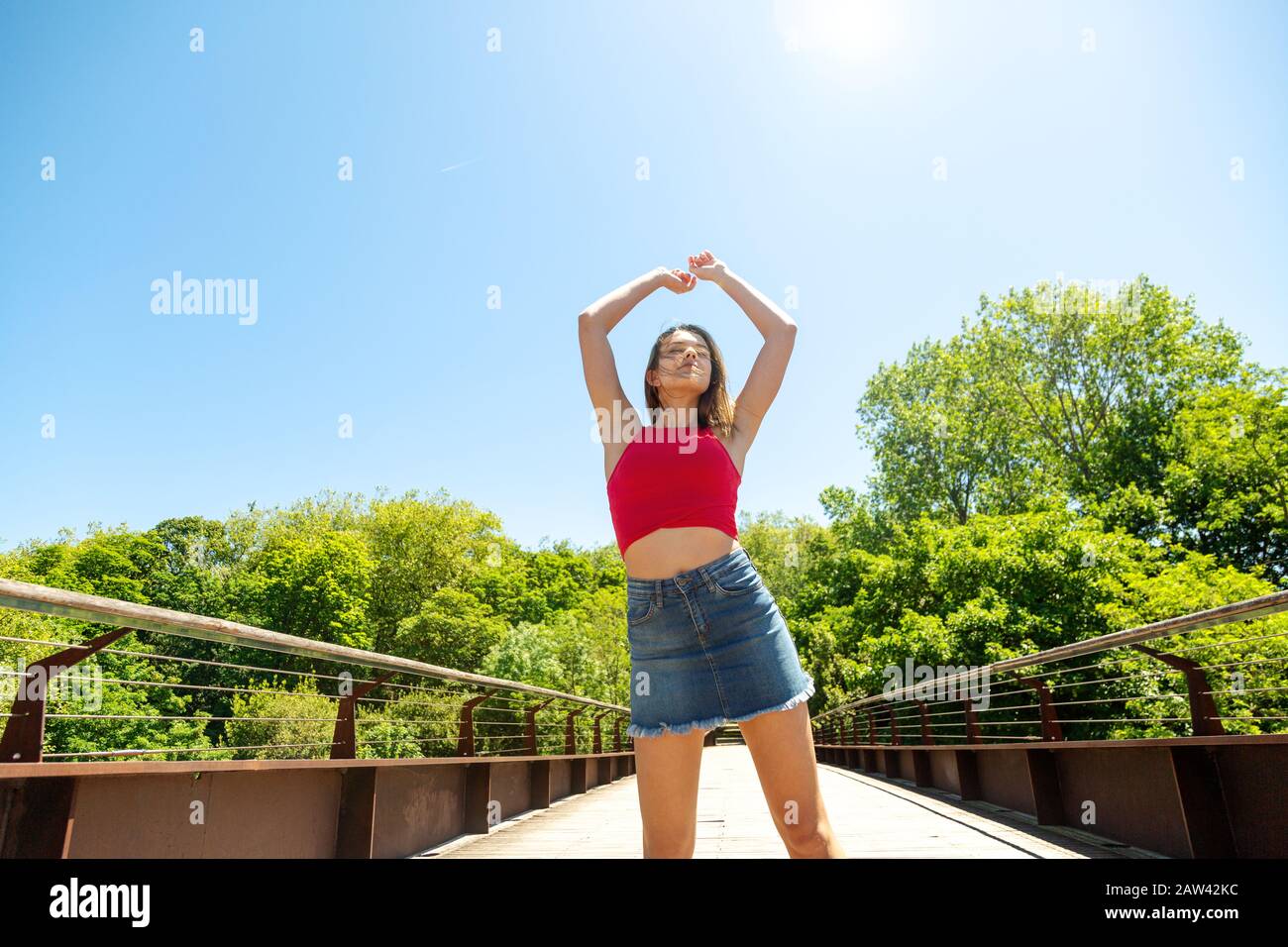 Happy young beautiful woman in red top and denim mini skirt with arms raised and eyes closed enjoying the sun on a bridge in a park in spring or summe Stock Photo