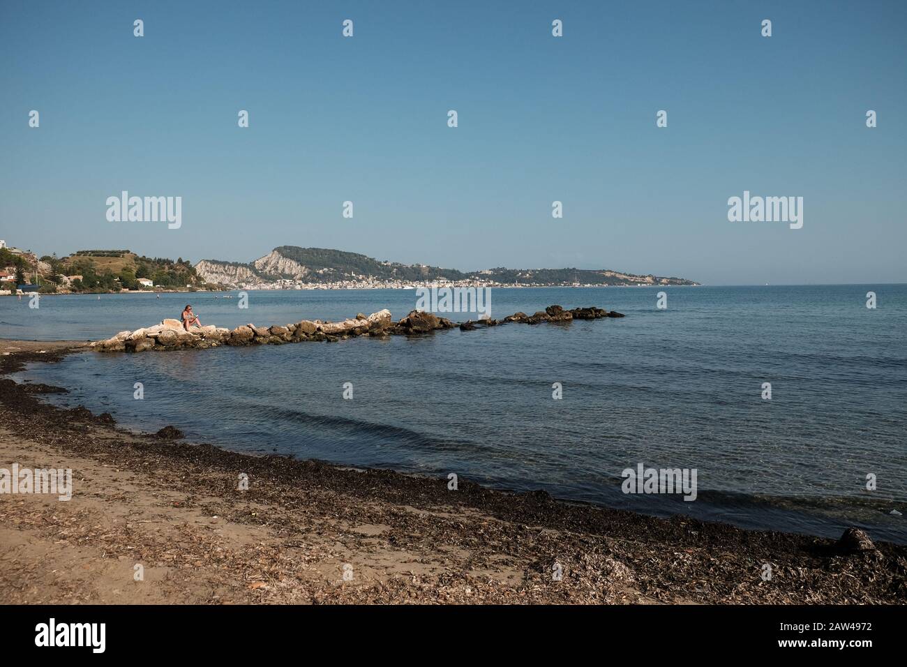A lone female is sat relaxing on the rocks reading a book on a warm sunny day at a empty beach on Zakynthos island Greece. Stock Photo