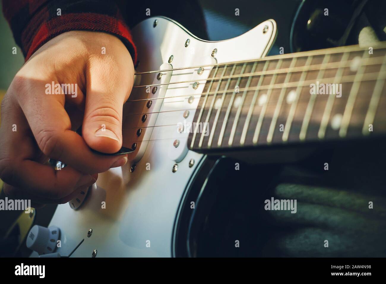 A musician in a red checked jacket plays with a mediator in his hand on the strings of an electronic black-and-white guitar, which is illuminated by a Stock Photo