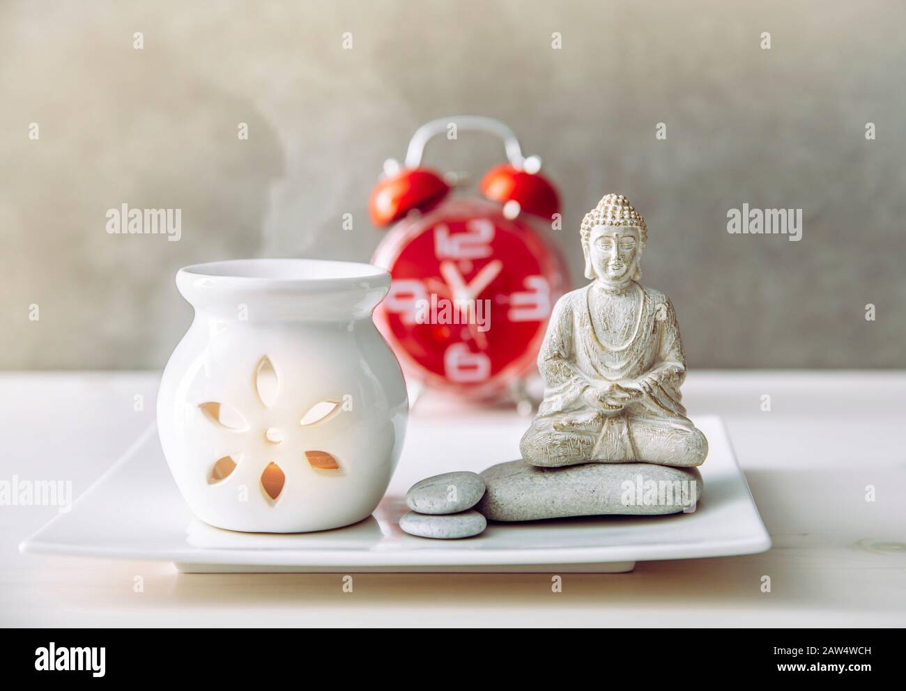 How to find balance between busy life and healthy mental health. Taking a break to sit and meditate concept. Buddha figure with aroma lamp and clock o Stock Photo