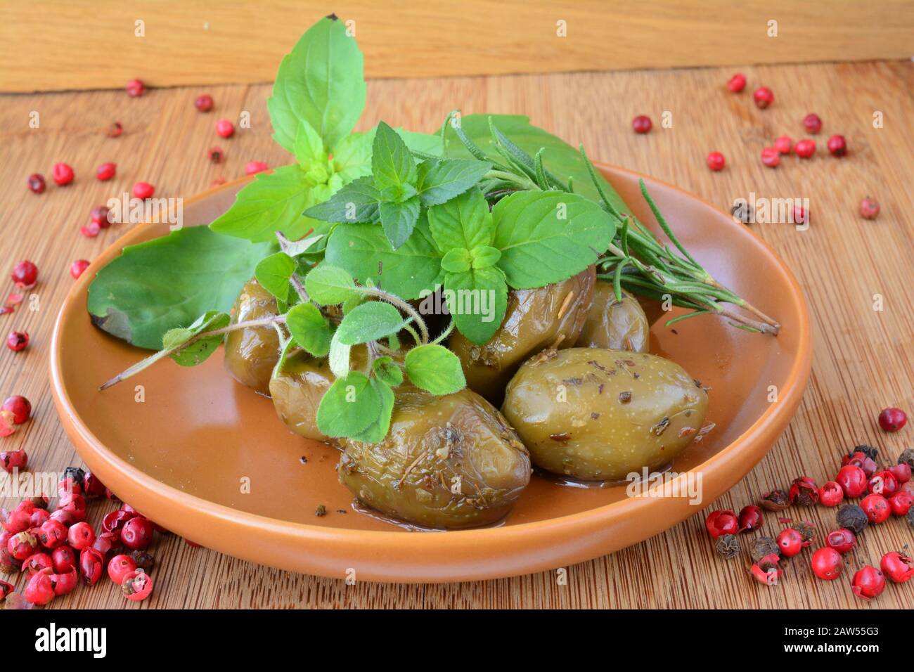 Aromatic green olives in rustic ceramic saucer, decorated with green basil, oregano, rosemary, peper and mojito mint leaves and red pepper, on wooden Stock Photo