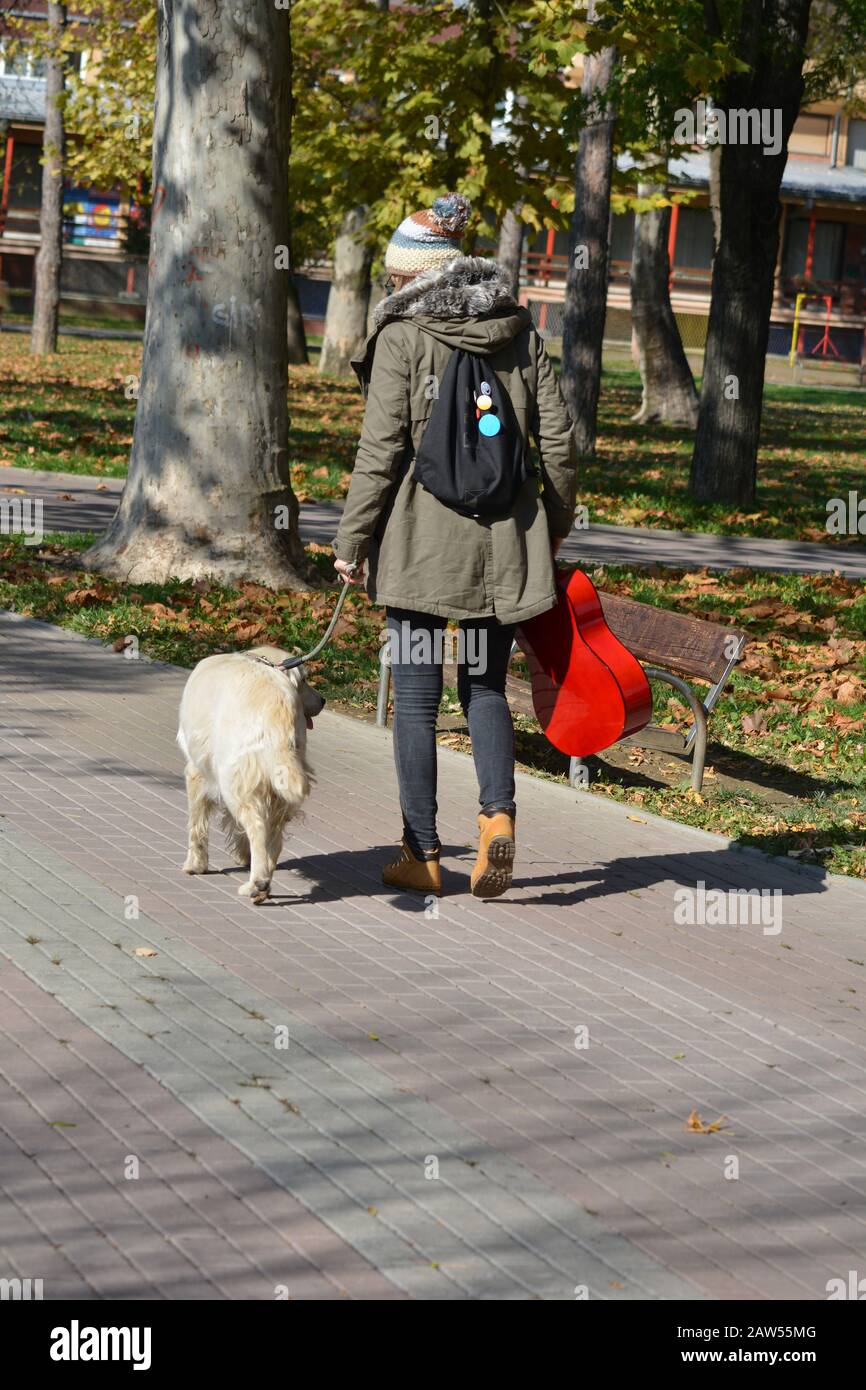 Young girl with wool cap, red guitar and small backpack walking with her golden retreiver dog in a park, view from behind Stock Photo