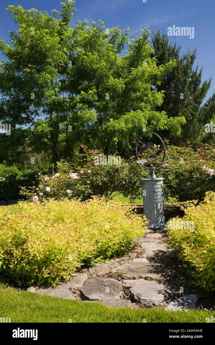 Flagstone path leading to a globe shaped sundial bordered by Spiraea japonica 'Gold Mound' - Spirea shrubs in backyard country garden in spring Stock Photo