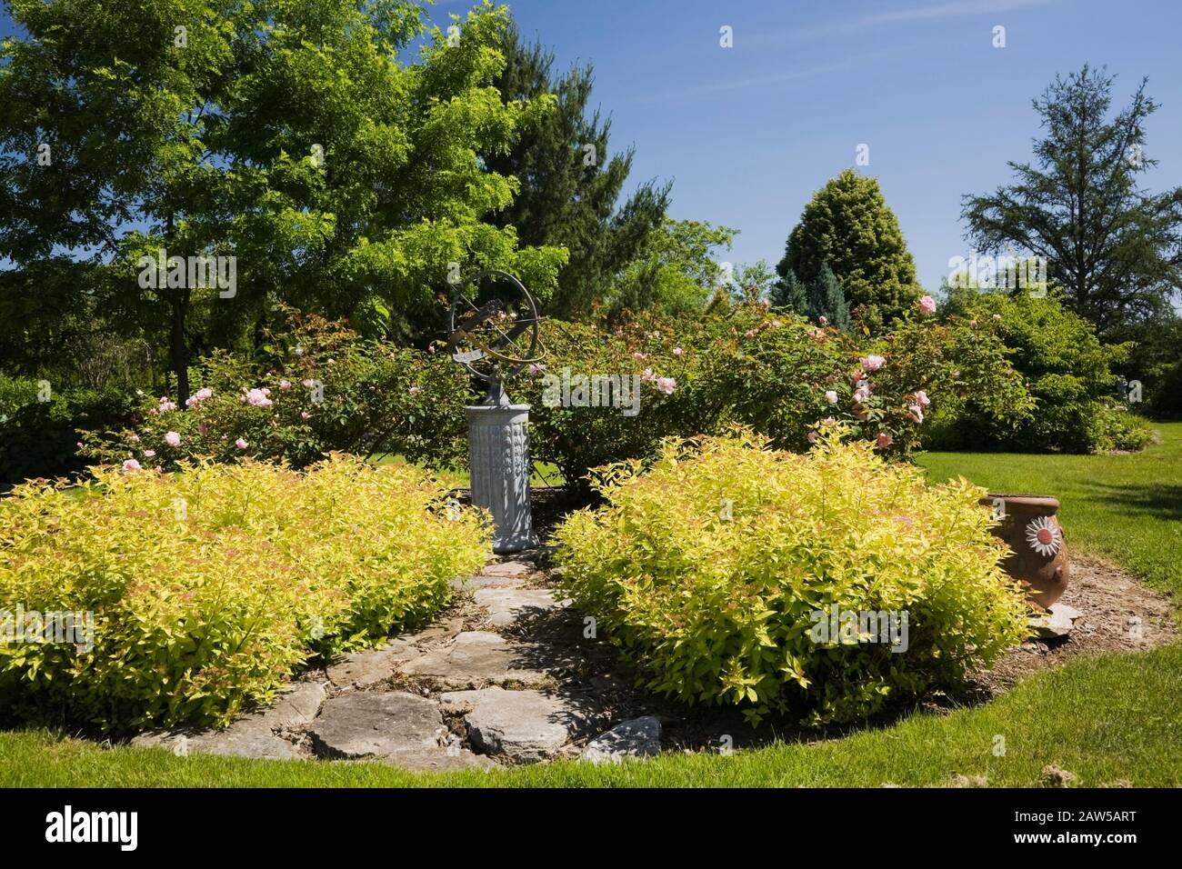 Flagstone path leading to a globe shaped sundial bordered by Spiraea japonica 'Gold Mound' - Spirea shrubs in backyard garden in spring Stock Photo