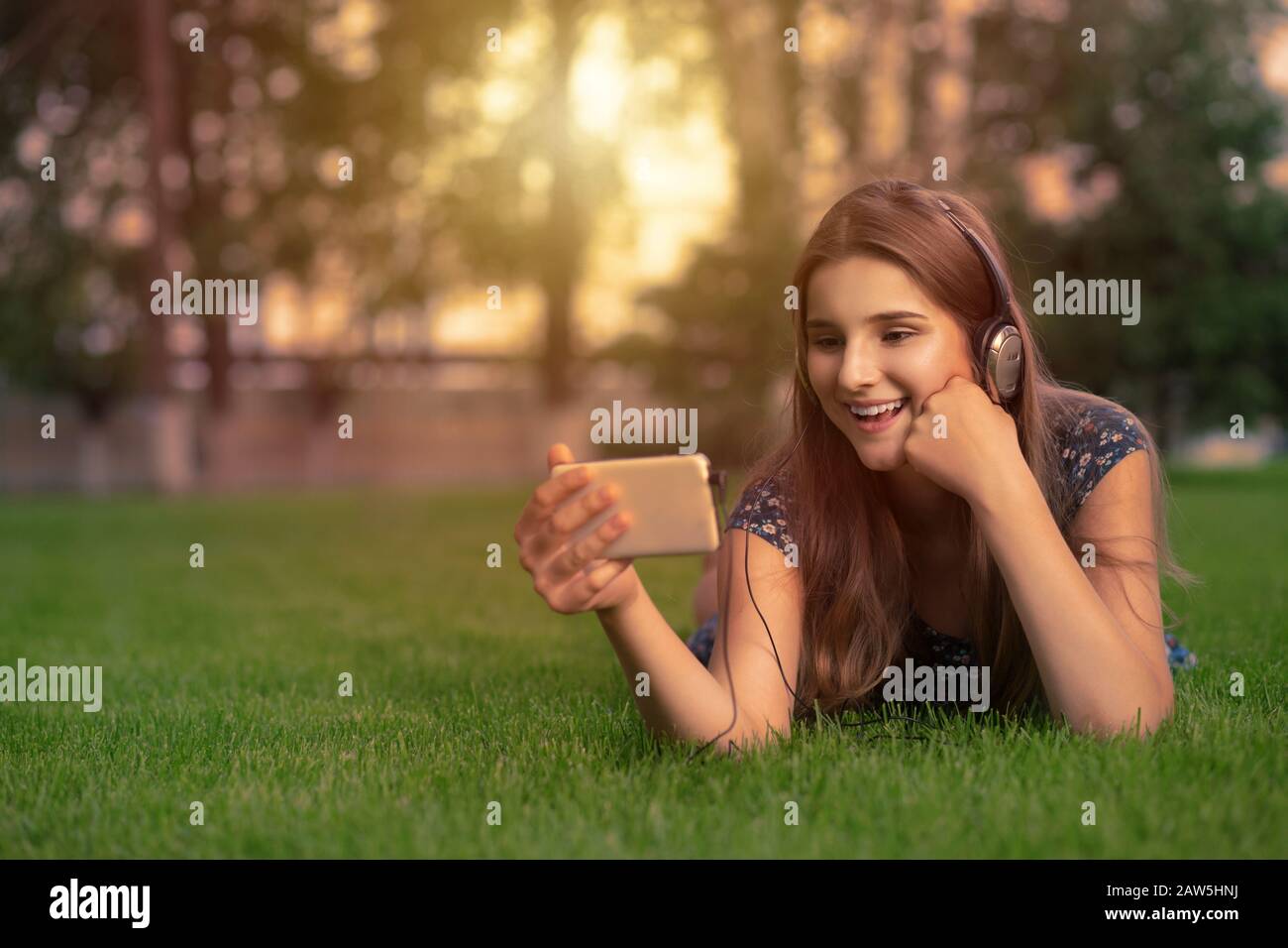 Happy young cute student woman with smartphone headset on head listening to music looking at watching game video film movie lying on green grass lawn Stock Photo