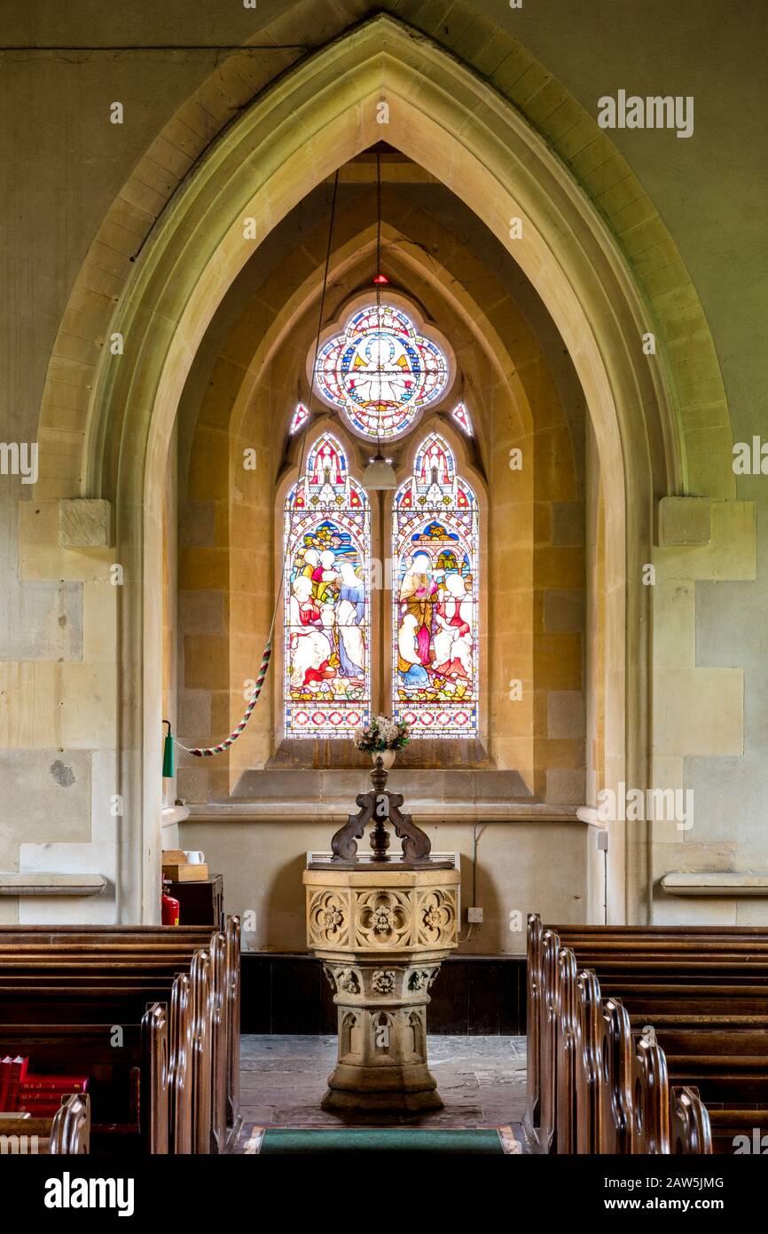 Stained glass window, baptism font and Gothic architecture of Saint Barnabas church in Snowshill, Gloucestershire, England, UK Stock Photo
