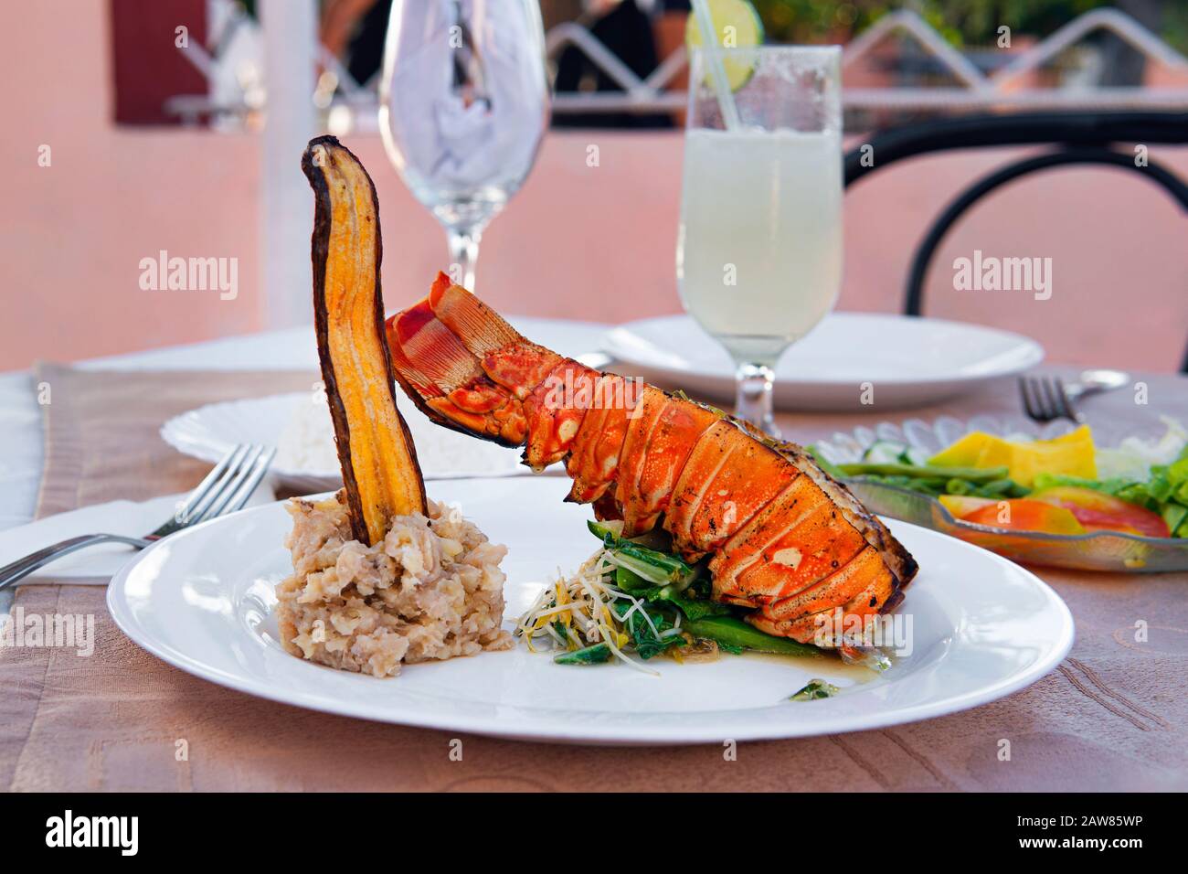 Grilled lobster is beautifully placed on a plate along with a side dish of mashed potatoes, fried banana, vegetables and rice and a glass of cocktail. Stock Photo