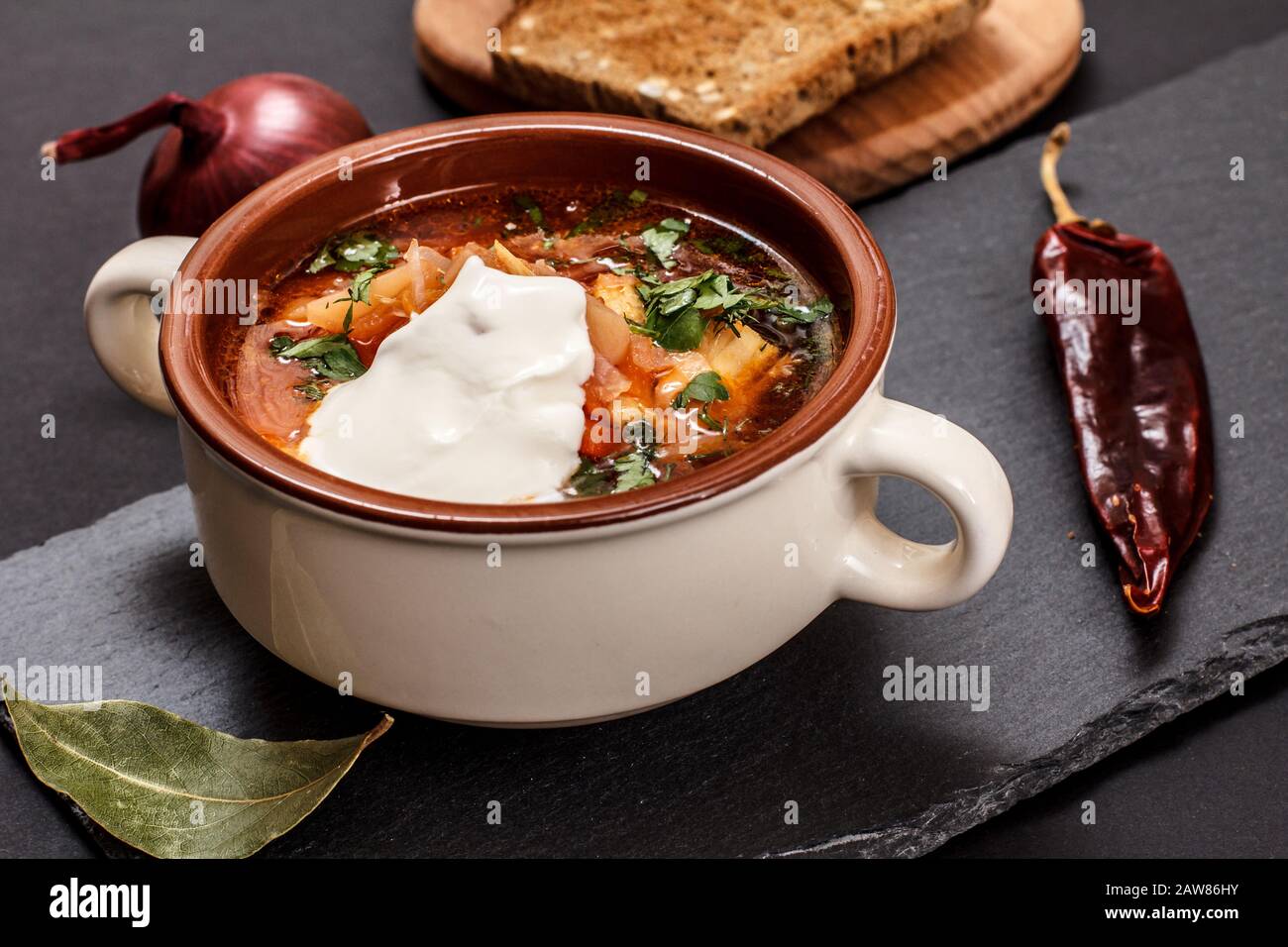 Ukrainian traditional borsch in porcelain bowl with sour cream, rye bread, onion and chilli pepper on stone board. Top view. Stock Photo