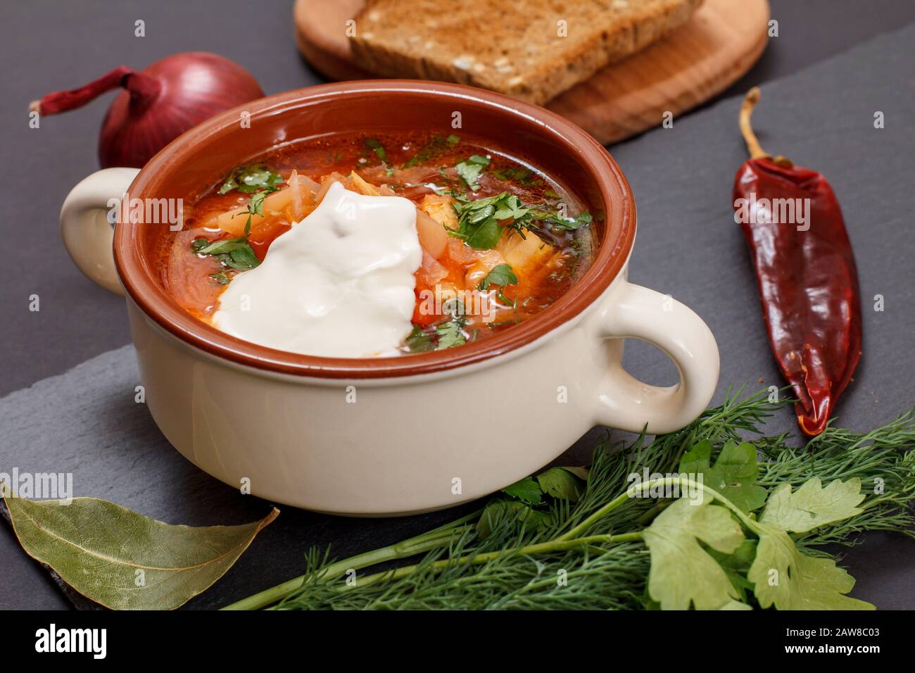 Ukrainian traditional borsch in porcelain bowl with sour cream, rye bread, onion, parsley and chilli pepper on stone board. Stock Photo