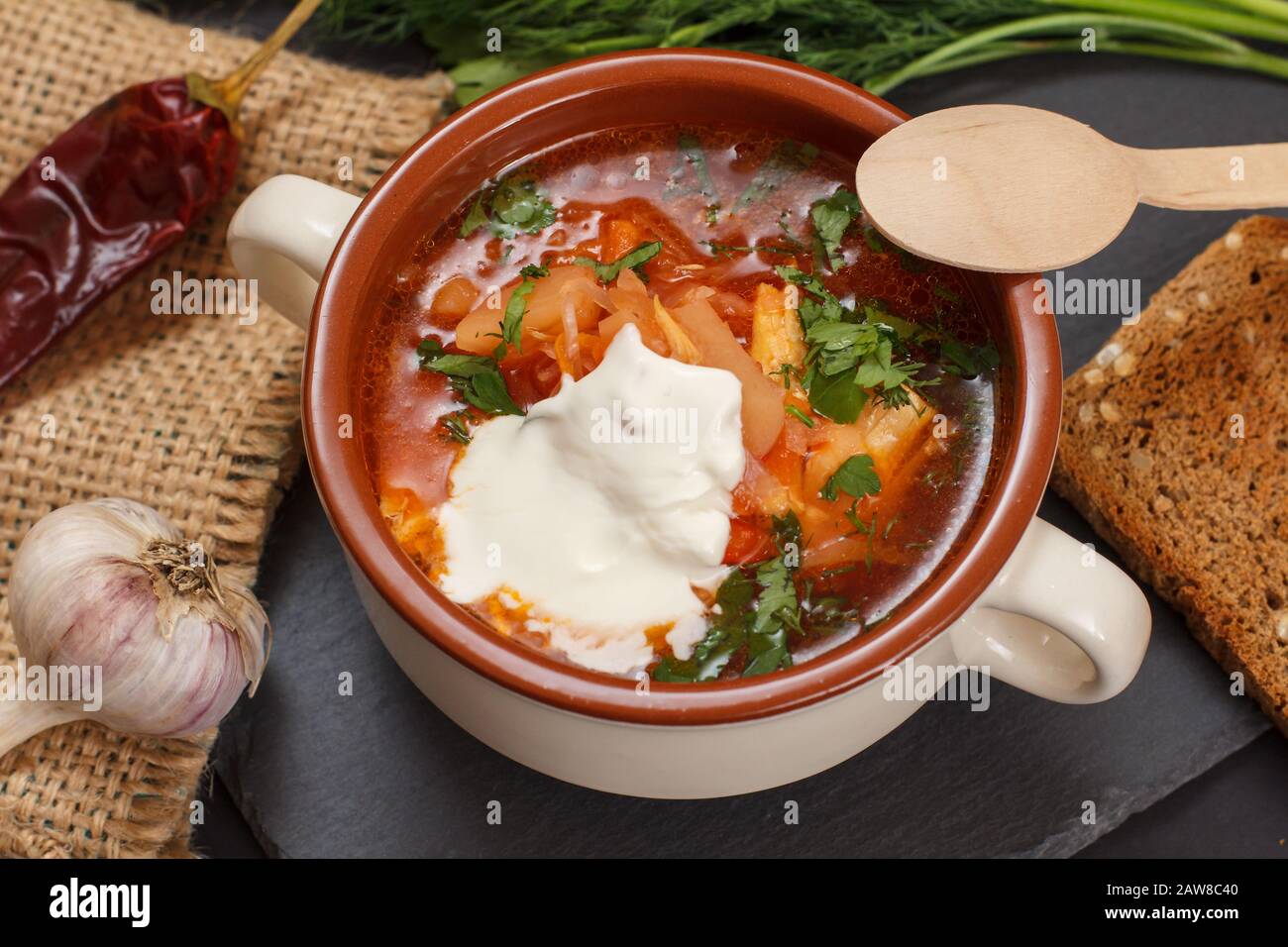 Ukrainian traditional borsch with sour cream in porcelain bowl with rye bread, parsley, garlic and chilli pepper on sackcloth and stone board. Top vie Stock Photo