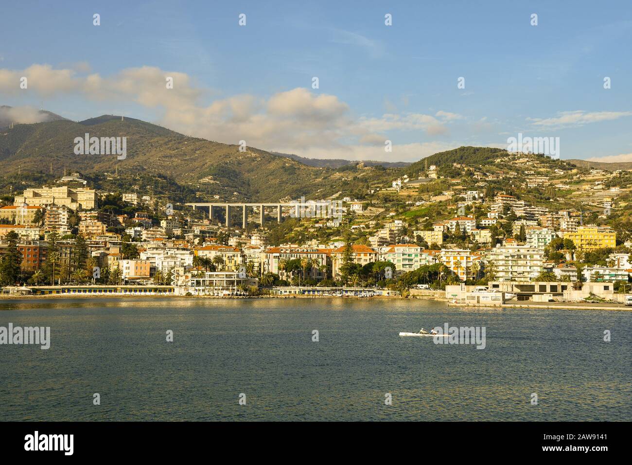 Scenic view of the coastal city from the sea with a passing canoe and the Ligurian Apennines mountains in a sunny day, Sanremo, Liguria, Italy Stock Photo