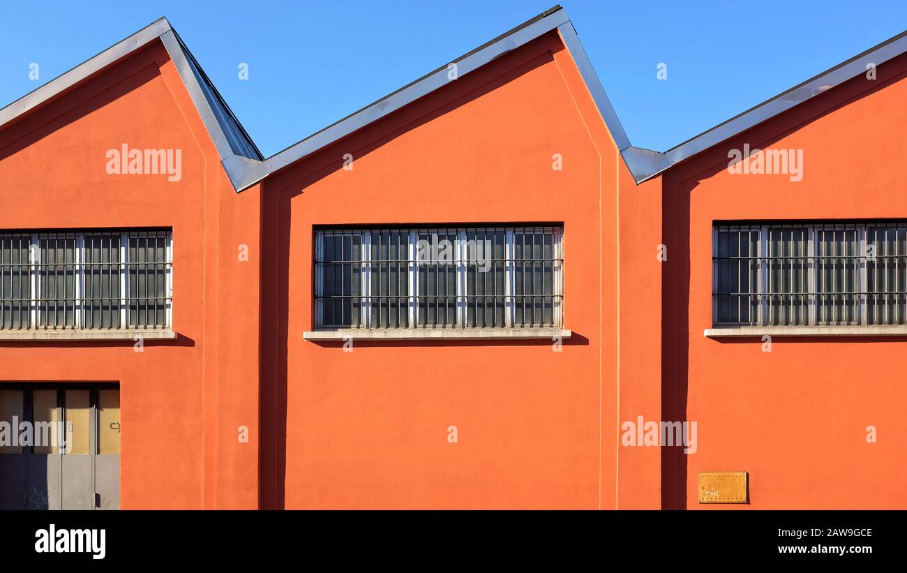 Old-fashioned factory shed with with a deep orange facade on a blue sky, zig-zag roof and windows in the southern suburbs of Milan, Italy Stock Photo