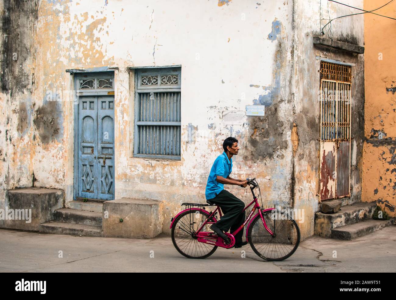 A man rides a bicycle past the decaying walls and quaint wooden doors and windows of old houses in the streets of the island of Diu in India. Stock Photo