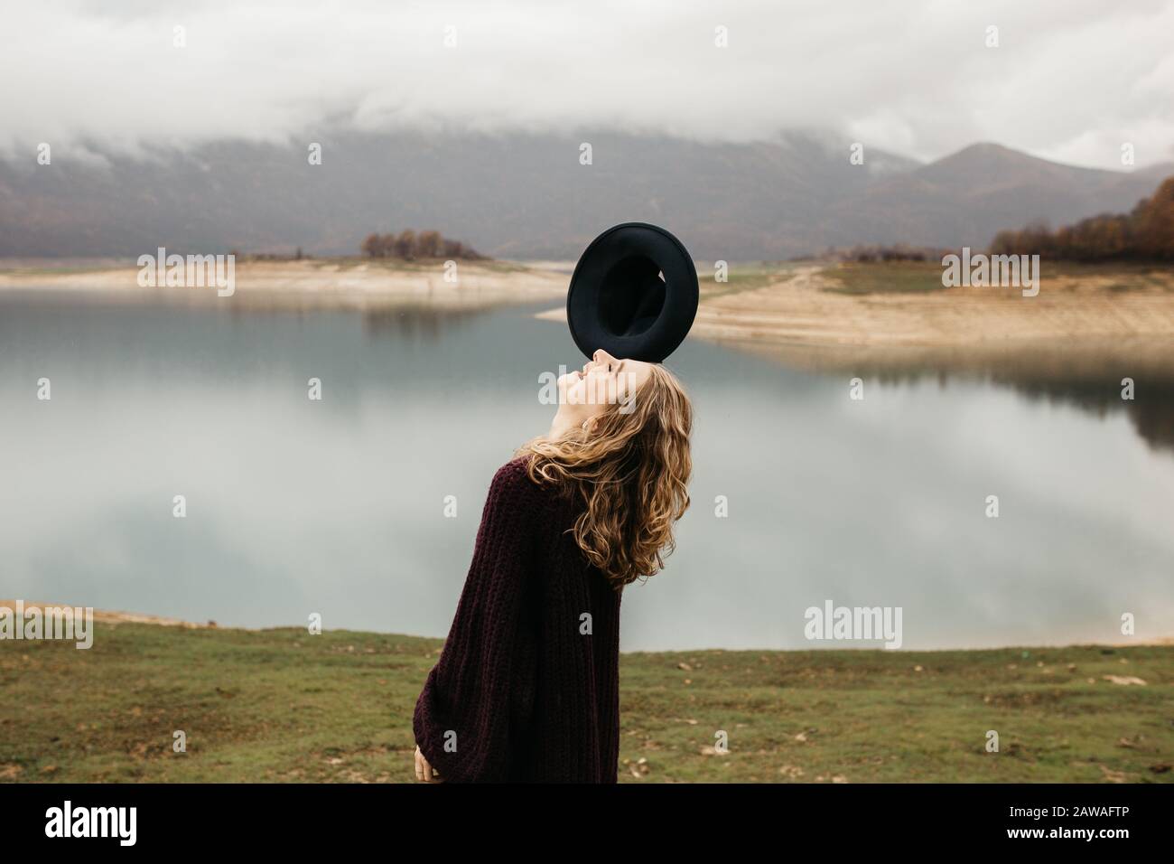 beautiful woman in purple sweater holding her hat and enjoying trip on the lake. Female tourist exploring lake. She is smiling and catching her hat. Stock Photo