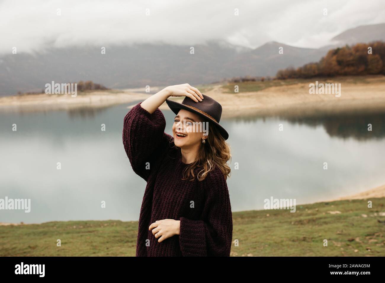 beautiful woman in purple sweater holding her hat and enjoying trip on the lake. Female tourist exploring lake. She is smiling and catching her hat. Stock Photo