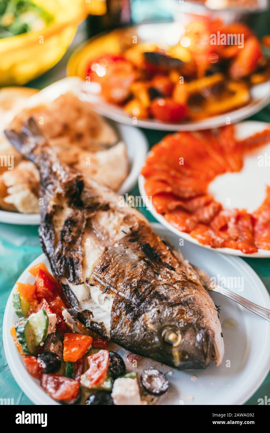 serving table with grilled fish, vegetable salad, salted fish and vegetables on plastic disposable plates. Selective focus macro shot with shallow DOF Stock Photo