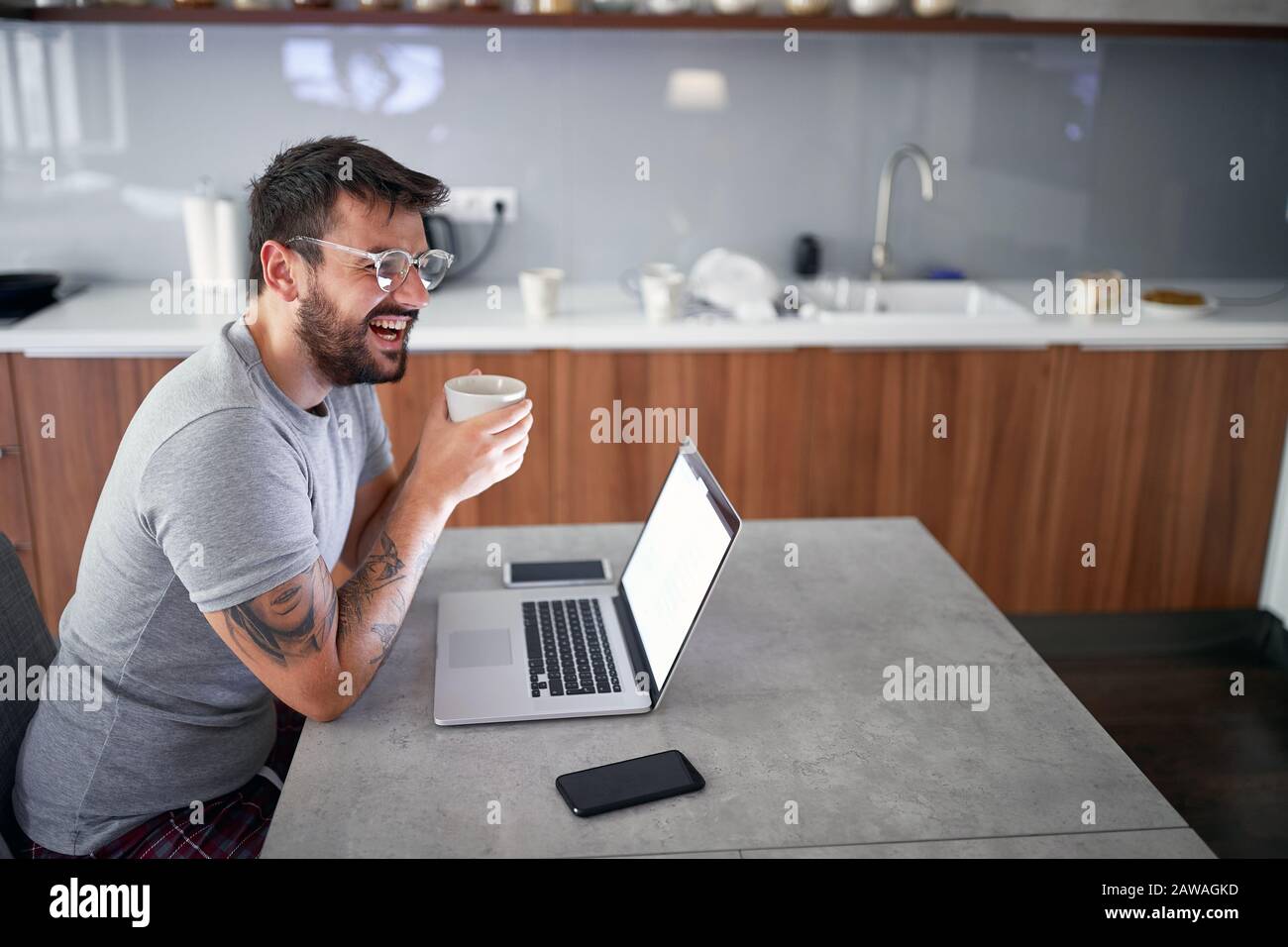 adult young male with beard wearing glasses,  smiling  in front of laptop on table, holding a cup. modern, casual, lifestyle, business concept Stock Photo