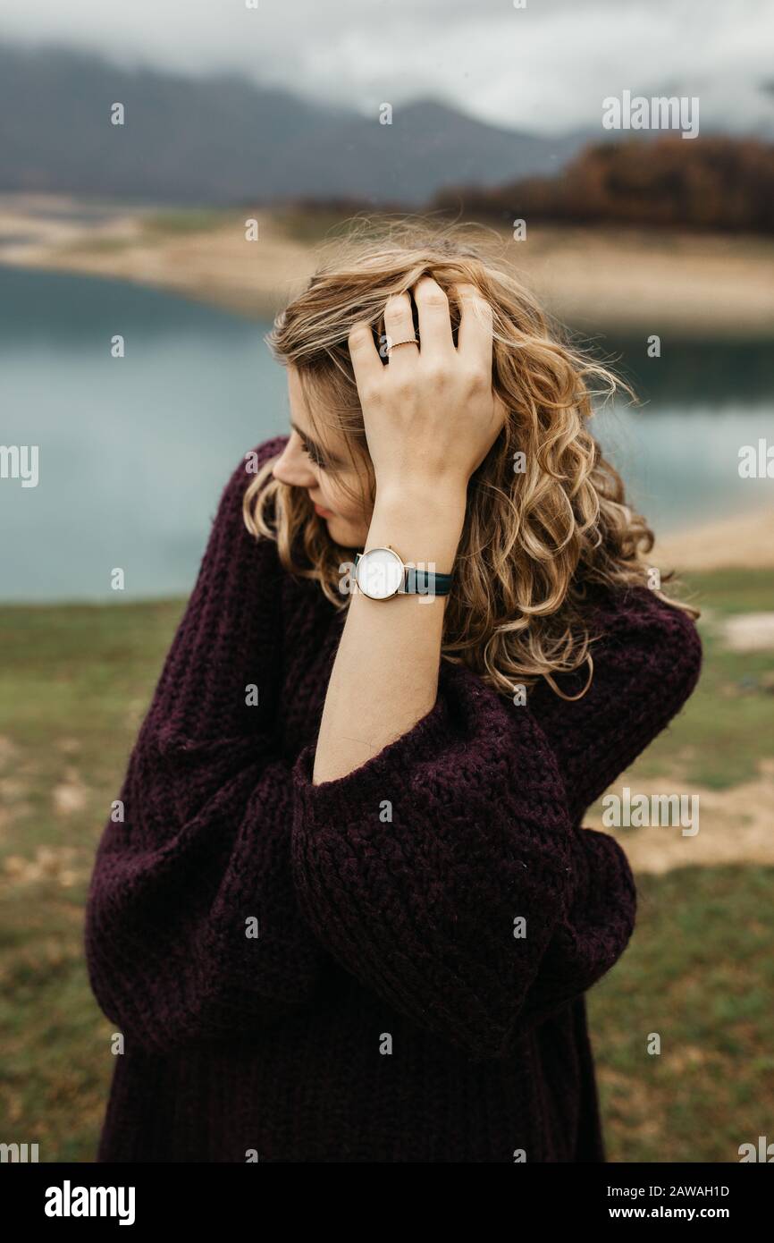 Outdoors portrait of young woman with curly hair wearing fashionable wrist watch. She is standing near lake. Stock Photo