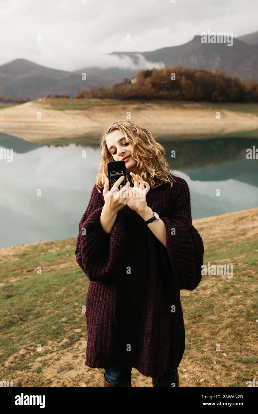 Beautiful woman with curly hair holding mobile phone and taking photos of lake on a cloudy day. She is texting on smartphone and taking selfie. Stock Photo