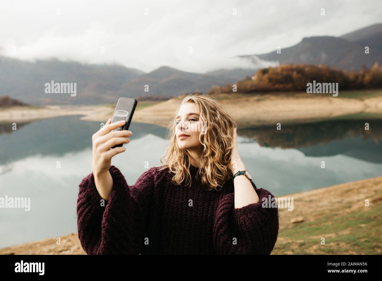 Beautiful woman with curly hair holding mobile phone and taking photos of lake on a cloudy day. She is texting on smartphone and taking selfie. Stock Photo