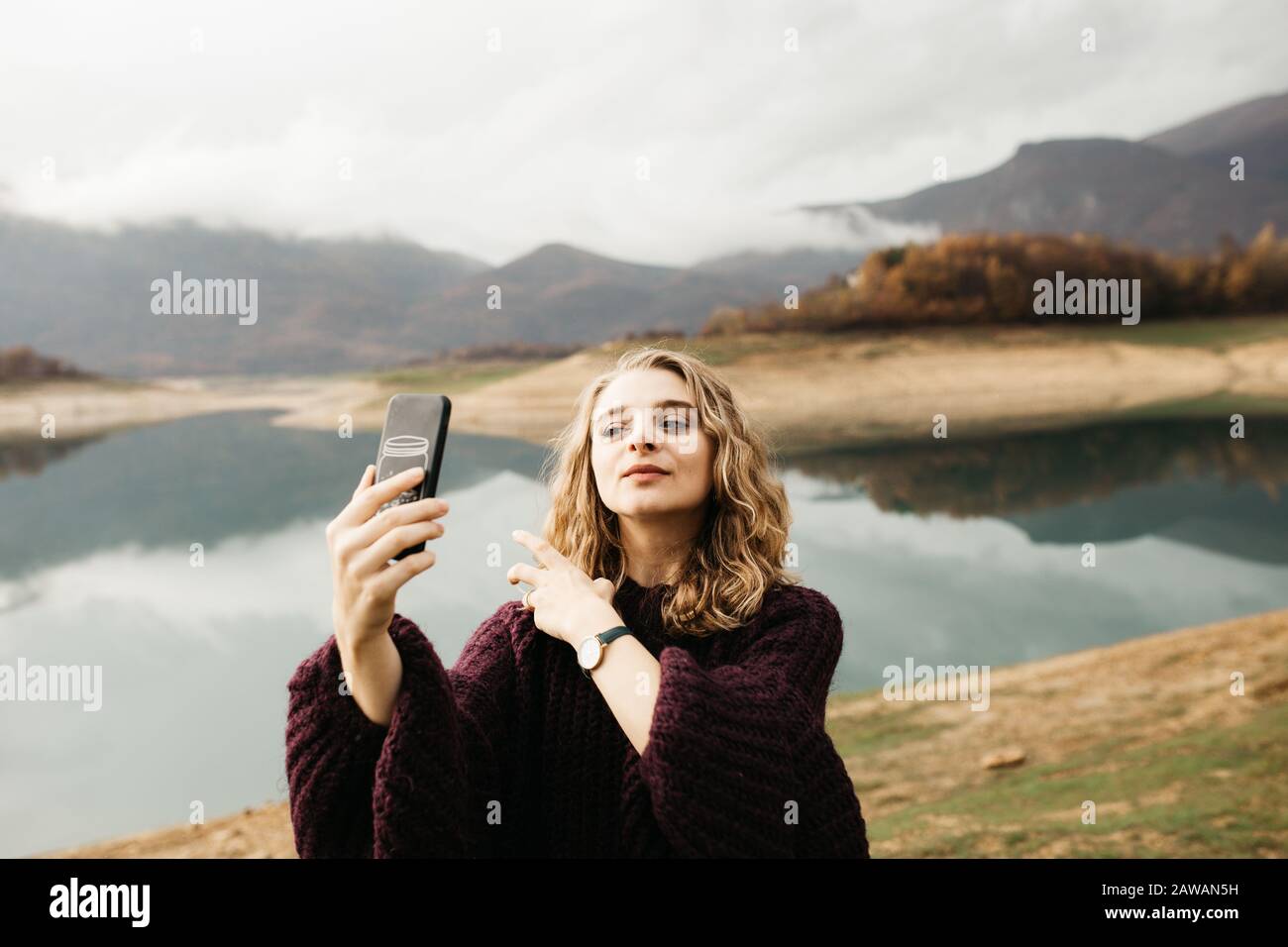 Beautiful woman with curly hair holding mobile phone and taking photos of lake on a cloudy day. She is texting on smartphone and taking selfie. Stock Photo