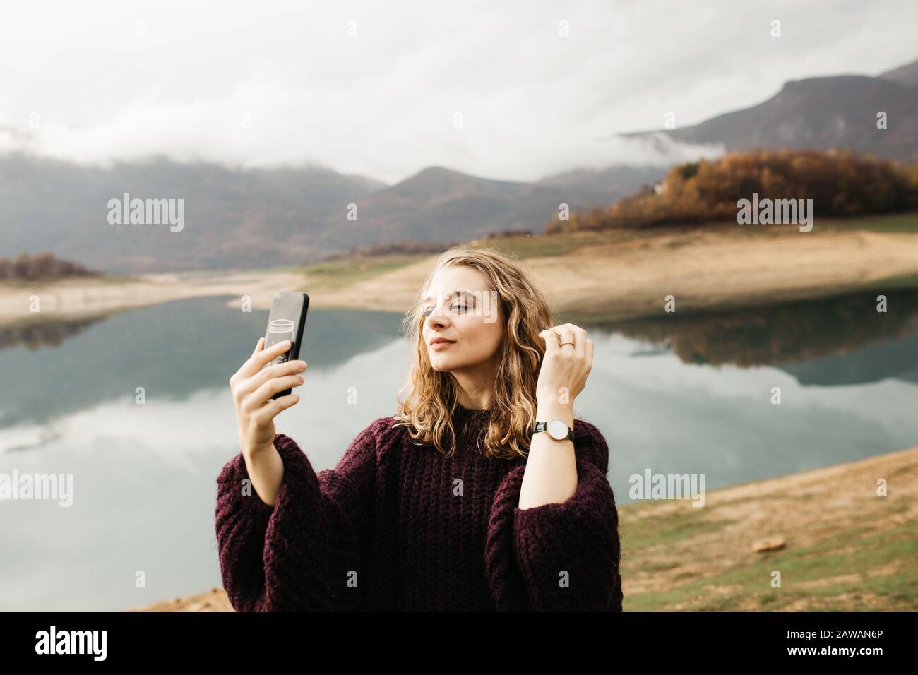 Beautiful woman with curly hair holding mobile phone and taking photos of lake on a cloudy day. She is texting on smartphone and taking selfie. Stock Photo