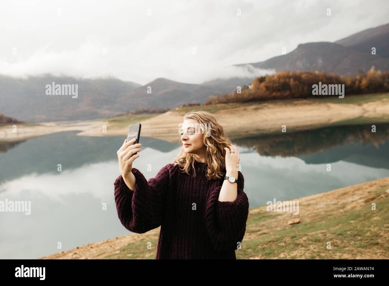 Beautiful woman with curly hair holding mobile phone and taking photos of lake on a cloudy day. She is texting on smartphone and taking selfie. Stock Photo