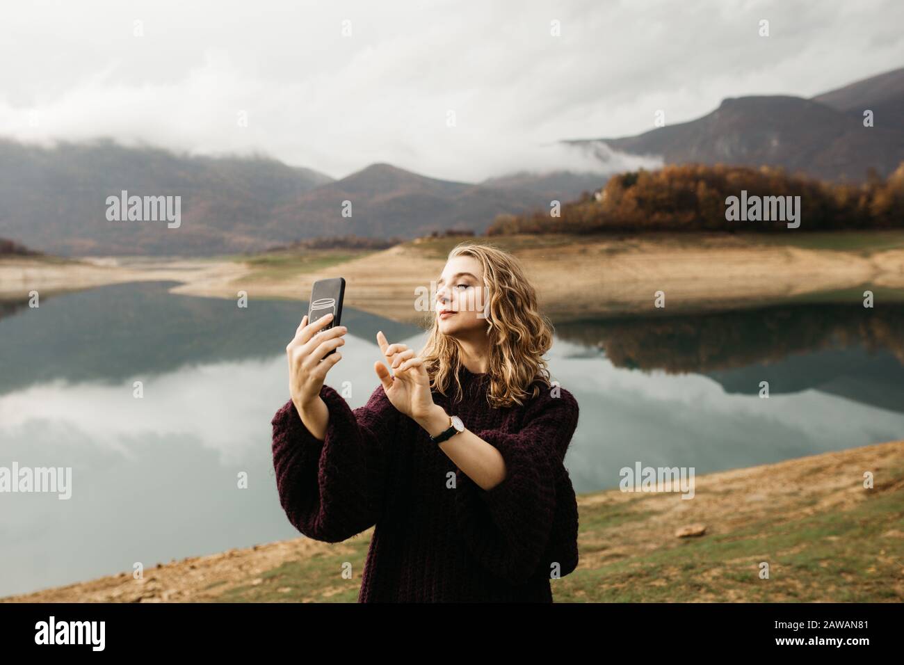 Beautiful woman with curly hair holding mobile phone and taking photos of lake on a cloudy day. She is texting on smartphone and taking selfie. Stock Photo