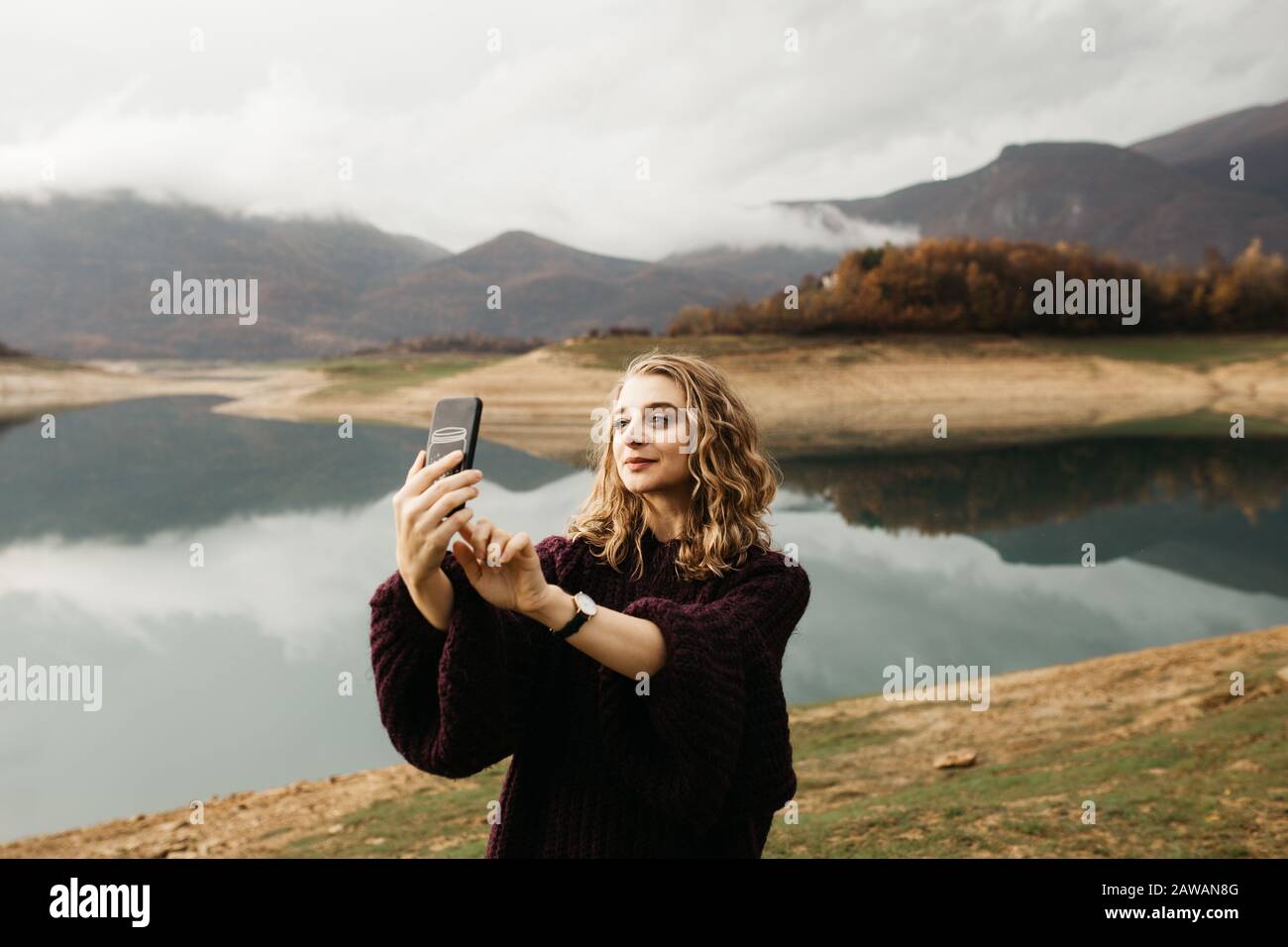 Beautiful woman with curly hair holding mobile phone and taking photos of lake on a cloudy day. She is texting on smartphone and taking selfie. Stock Photo