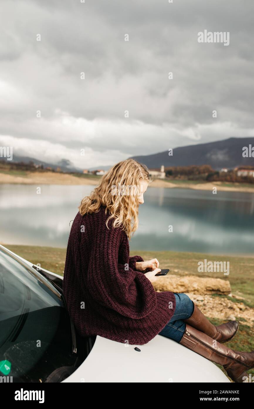 Beautiful woman with curly hair, sitting on the car, holding mobile phone and taking photos of lake on a cloudy day. She is texting on smartphone and Stock Photo