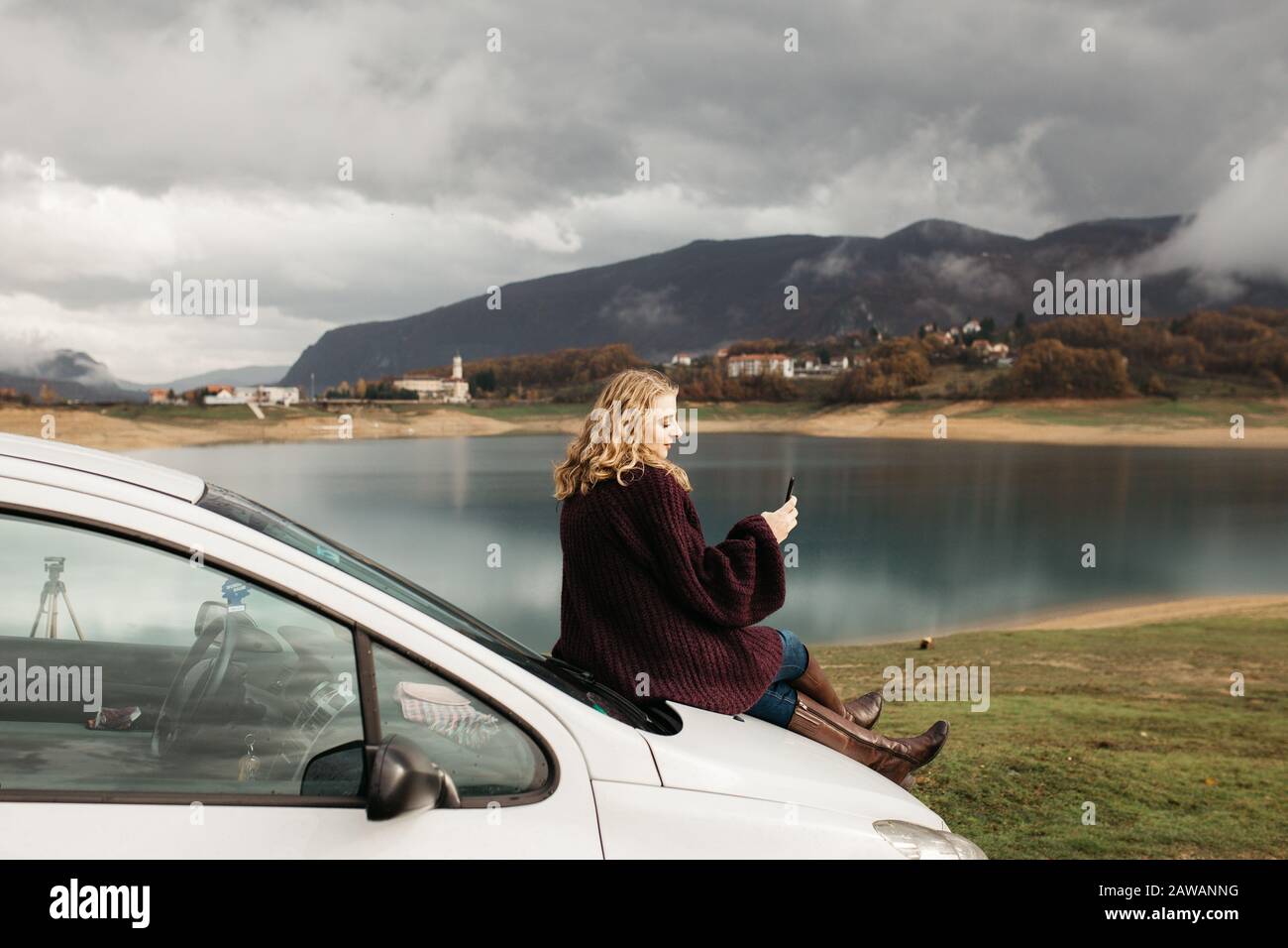 Beautiful woman with curly hair, sitting on the car, holding mobile phone and taking photos of lake on a cloudy day. She is texting on smartphone and Stock Photo