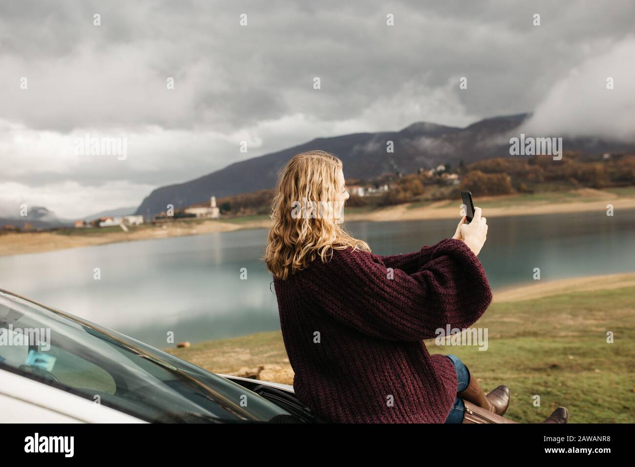 Beautiful woman with curly hair, sitting on the car, holding mobile phone and taking photos of lake on a cloudy day. She is texting on smartphone and Stock Photo