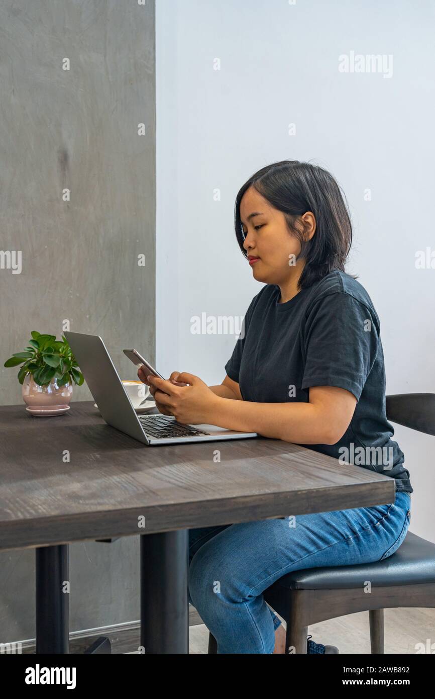 Asian female freelancer using smartphone while working on laptop Stock Photo