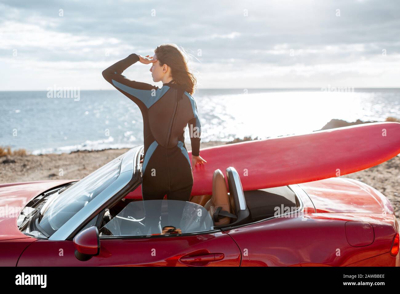 Woman enjoying beautiful landscapes while standing on her sports car with a surfboard near the ocean. Carefree lifestyle and active sports concept Stock Photo