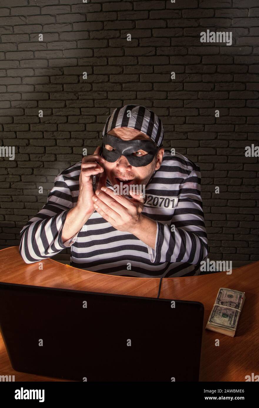 Comic character prisoner telephone con man sitting at a laptop in a striped robe and a hat in a black mask and cheating on the phone Stock Photo