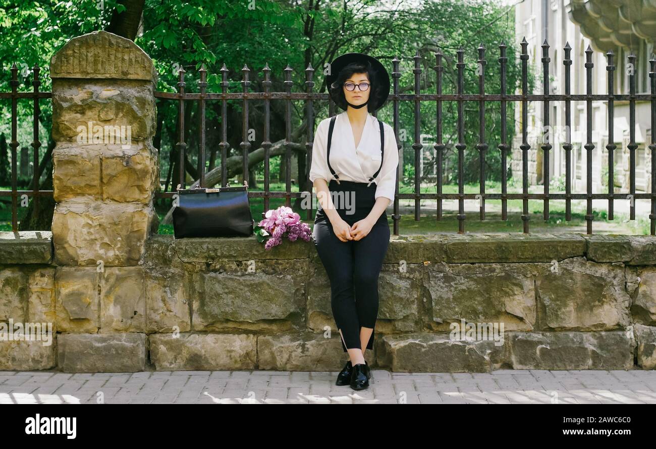 A beautiful girl with hat on her head and in glasses sita on the stone fence and looking to the sides. Beautiful summer day. Stock Photo