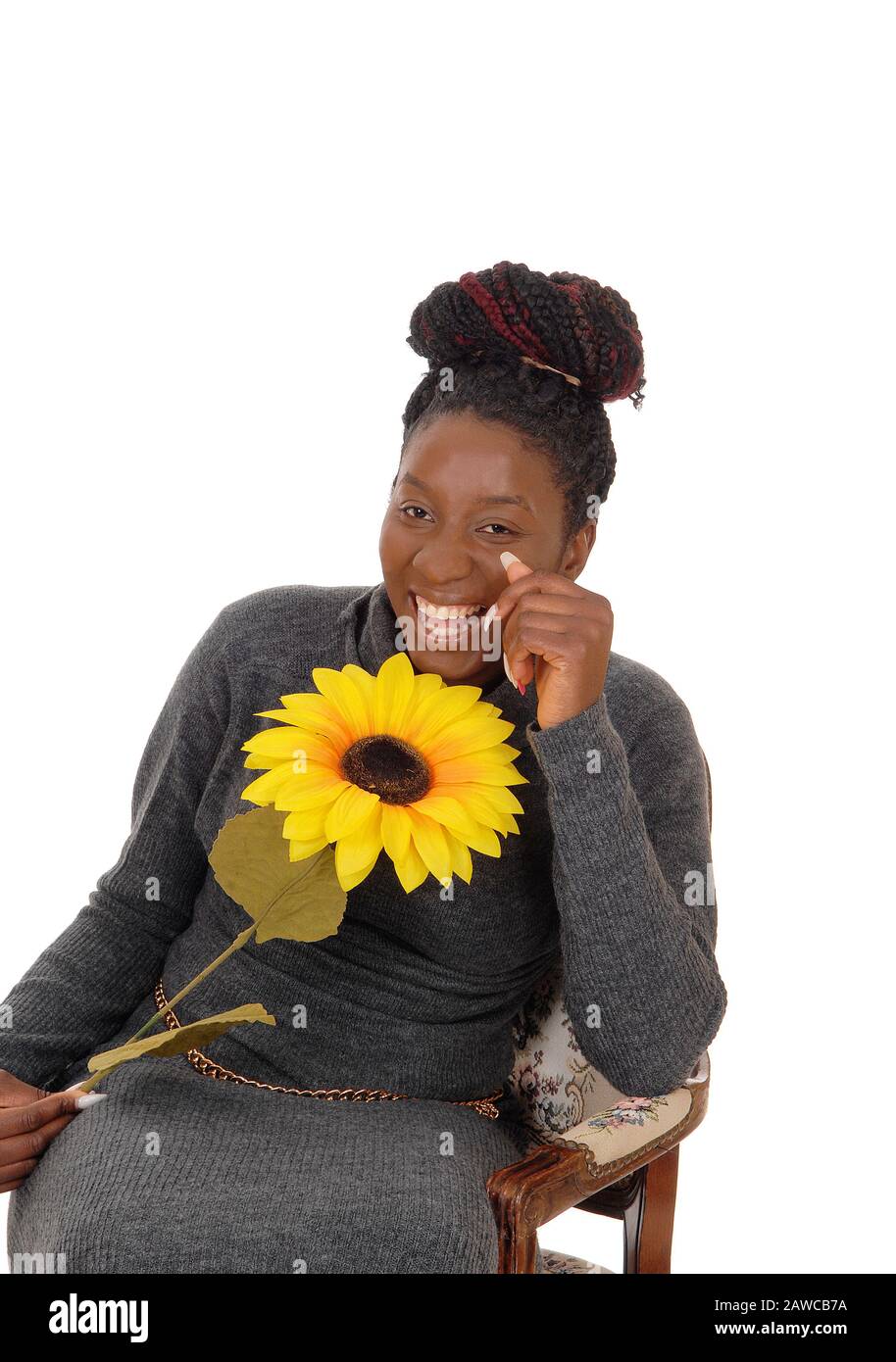 A beautiful young African American woman sitting smiling in a chair holding a yellow sunflower, isolated for white background Stock Photo