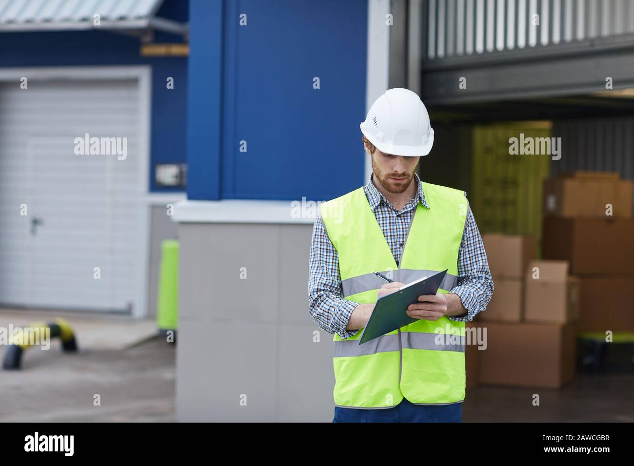 Young man in protective workwear standing and making notes in document while delivering load he working outdoors Stock Photo